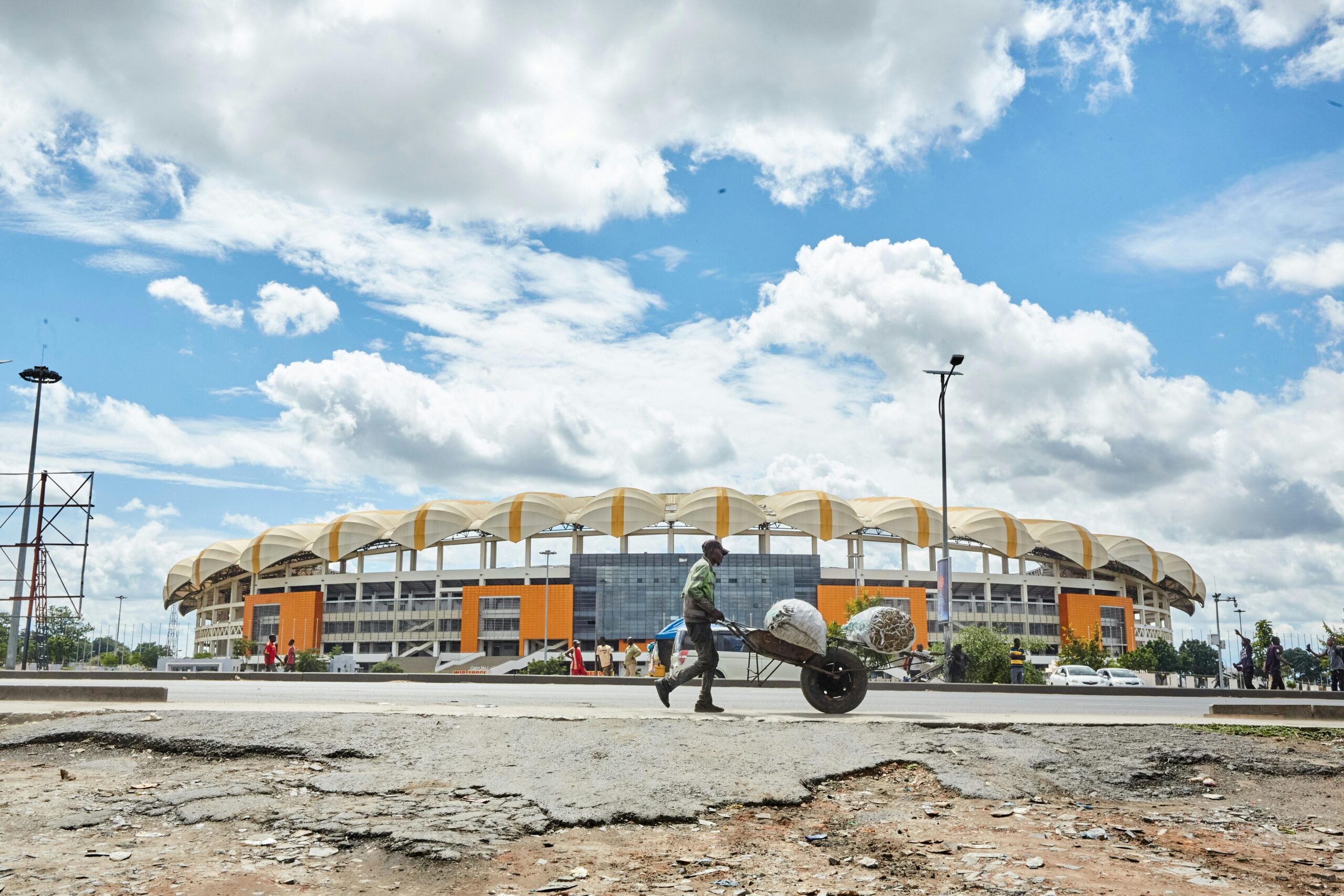 A person with a wheelbarrow in front of Lusaka Stadium under a blue sky with clouds.